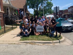 Group of 22 FleishmanHillard colleagues dressed in casual summer attire pose for a group photo in a neighborhood with a sign that reads, "We're all in this together."
